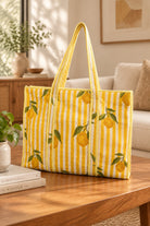A yellow and white striped tote bag with a lemon print sits on a wooden table in a bright, modern living room setting.
