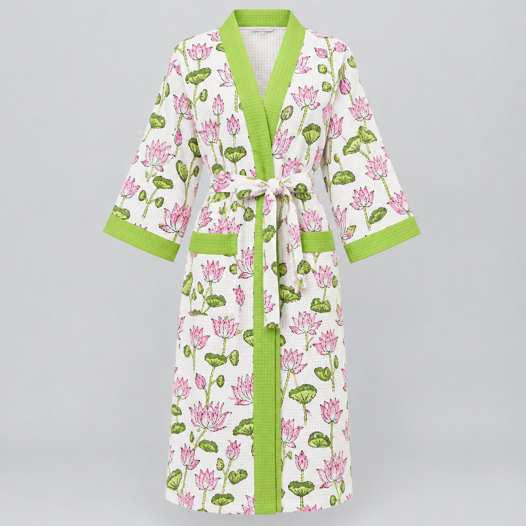 A white cotton kimono-style robe with a pink and green lotus print, featuring lime green trim and a matching tie belt, hanging on a wooden hanger against a background of green foliage.