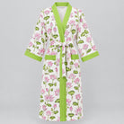 A white cotton kimono-style robe with a pink and green lotus print, featuring lime green trim and a matching tie belt, hanging on a wooden hanger against a background of green foliage.