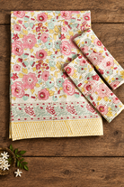 A floral cotton table linen set including a placemat and matching napkins, featuring pink and red flowers with yellow and green accents on a cream background, styled on a rustic wooden table.