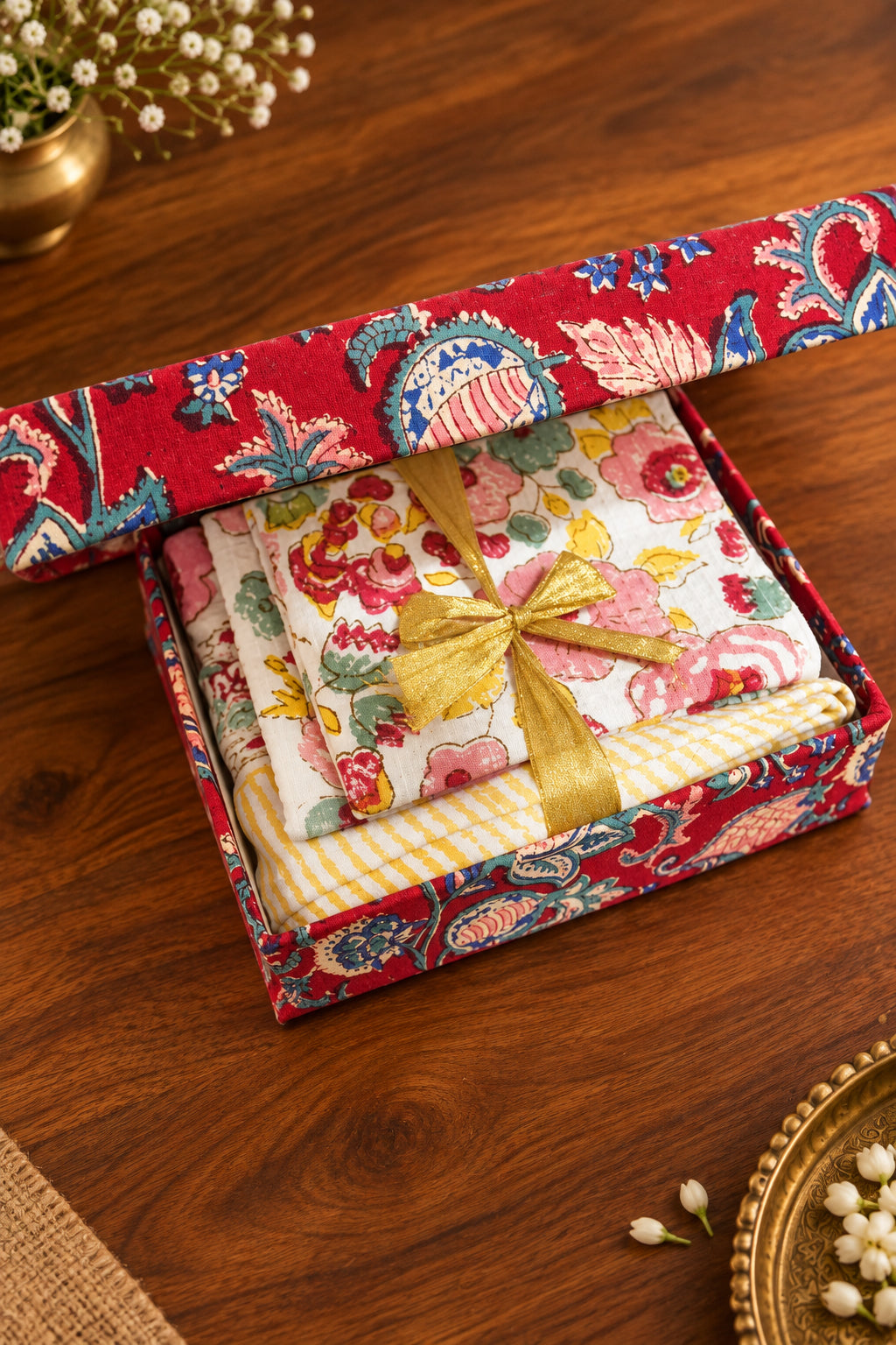 A gift box with a red paisley patterned lid and base, containing folded floral fabric pieces in pink, yellow, and white, tied with a gold ribbon, resting on a dark wooden surface.