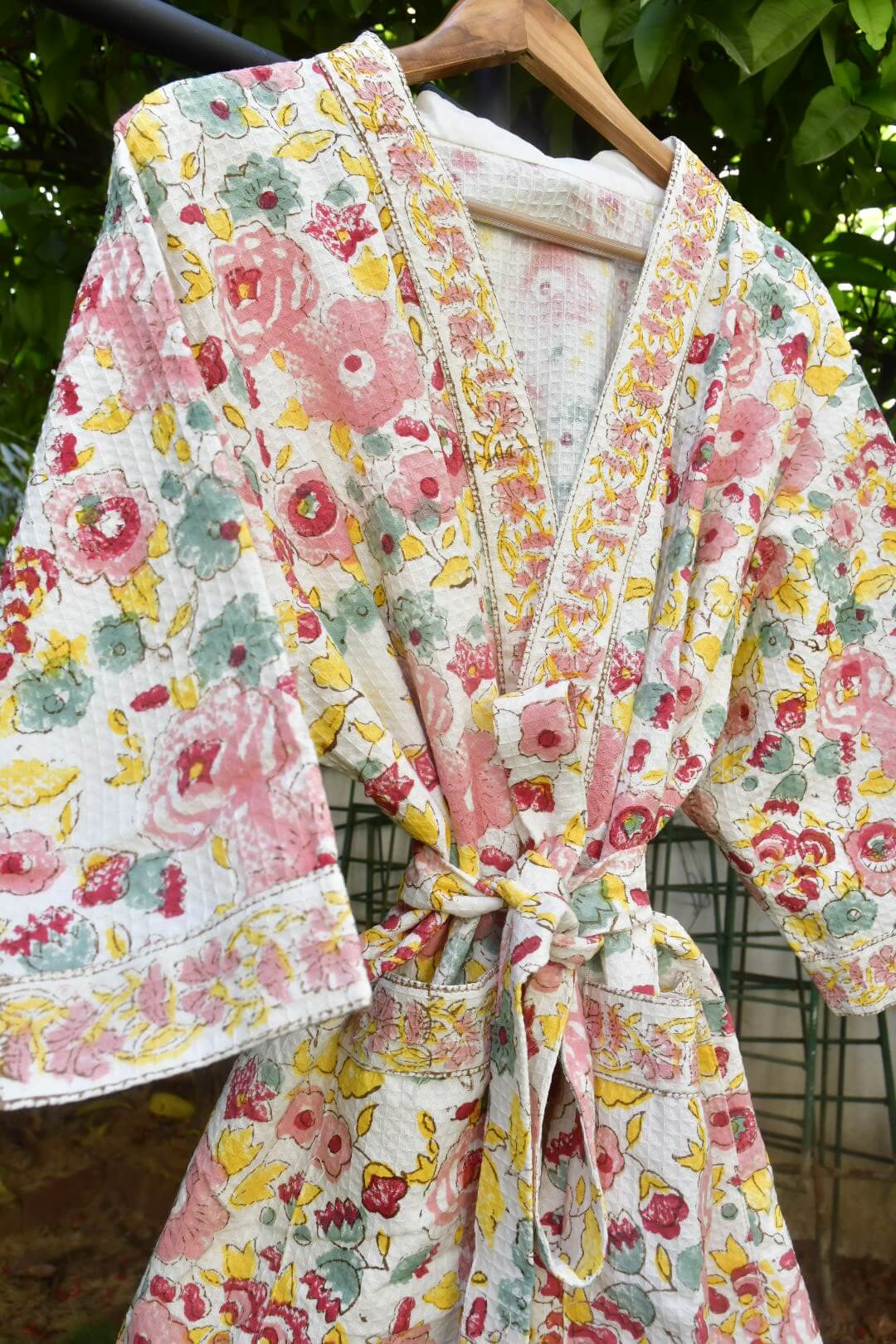 A floral print cotton robe with a tie waist, hanging on a wooden hanger against a background of green foliage.