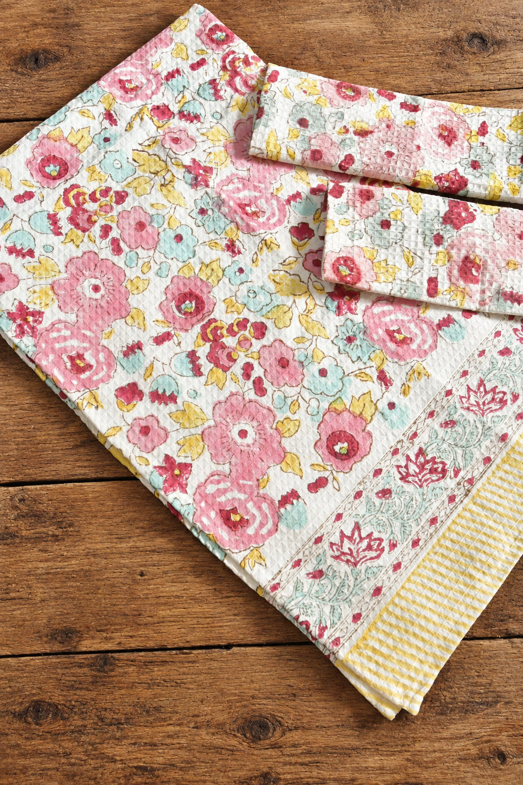 A folded cotton kitchen towel with a colorful floral print and a yellow striped border, displayed on a rustic wooden surface.