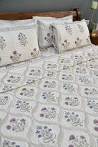 A quilted bedspread and matching pillow shams featuring a cream background with embroidered floral motifs in purple, blue, and green, styled on a bed with a wooden headboard.