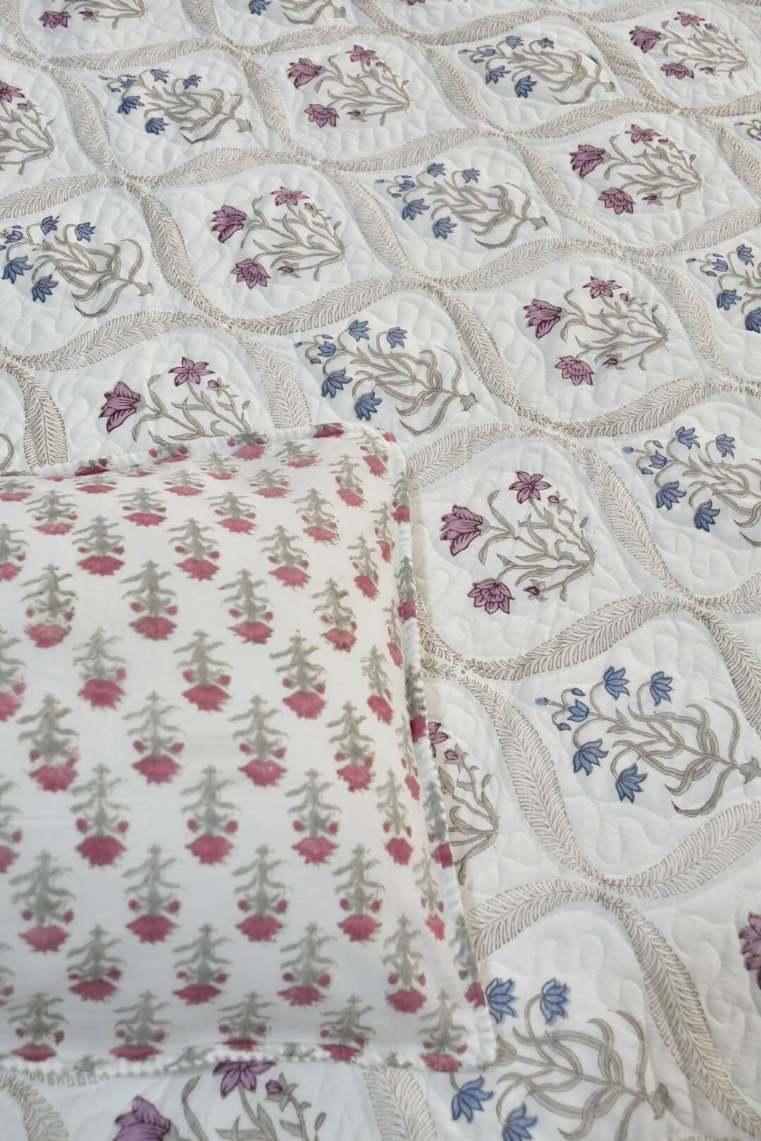 A close-up of a quilted textile with a repeating floral pattern in pink, blue, and gray on an off-white background, with a matching folded pillow or cushion shown in the lower left.