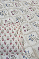 A close-up of a quilted textile with a repeating floral pattern in pink, blue, and gray on an off-white background, with a matching folded pillow or cushion shown in the lower left.