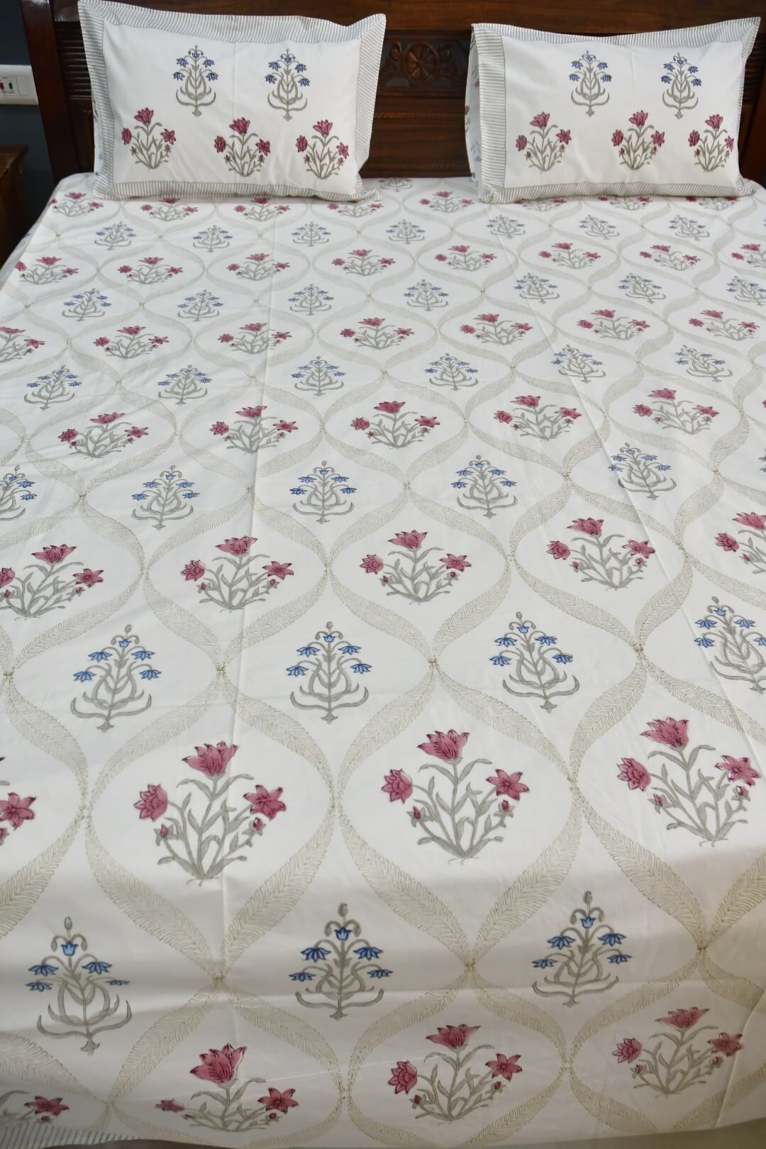 A white cotton bedsheet with matching pillowcases, featuring a repeating beige trellis pattern and floral motifs in pink, blue, and gray, styled on a bed with a dark wooden headboard.
