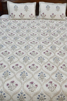 An quilted bedspread with matching pillowcases & bedsheet, featuring a cream background with a repeating geometric trellis pattern and floral motifs in purple, blue, and beige.