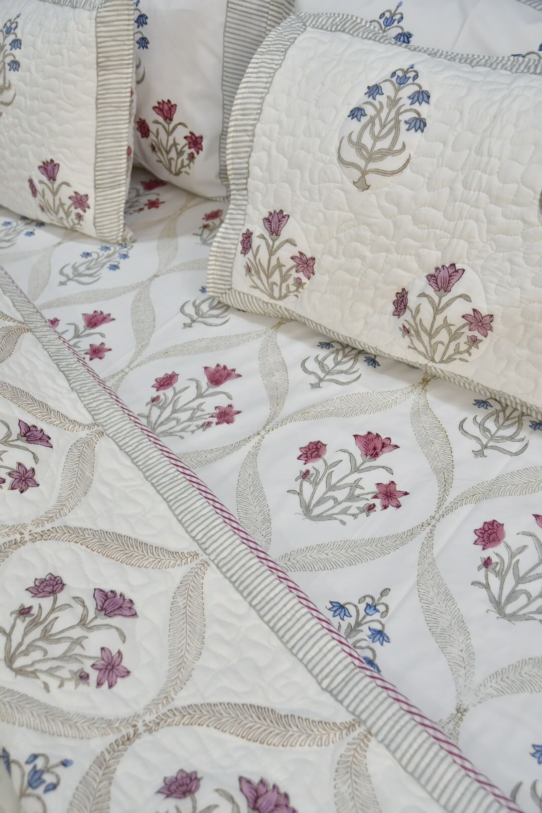 A close-up of a quilted bedding set featuring cream-colored fabric with embroidered floral motifs in pink, purple, and blue, accented by striped borders.