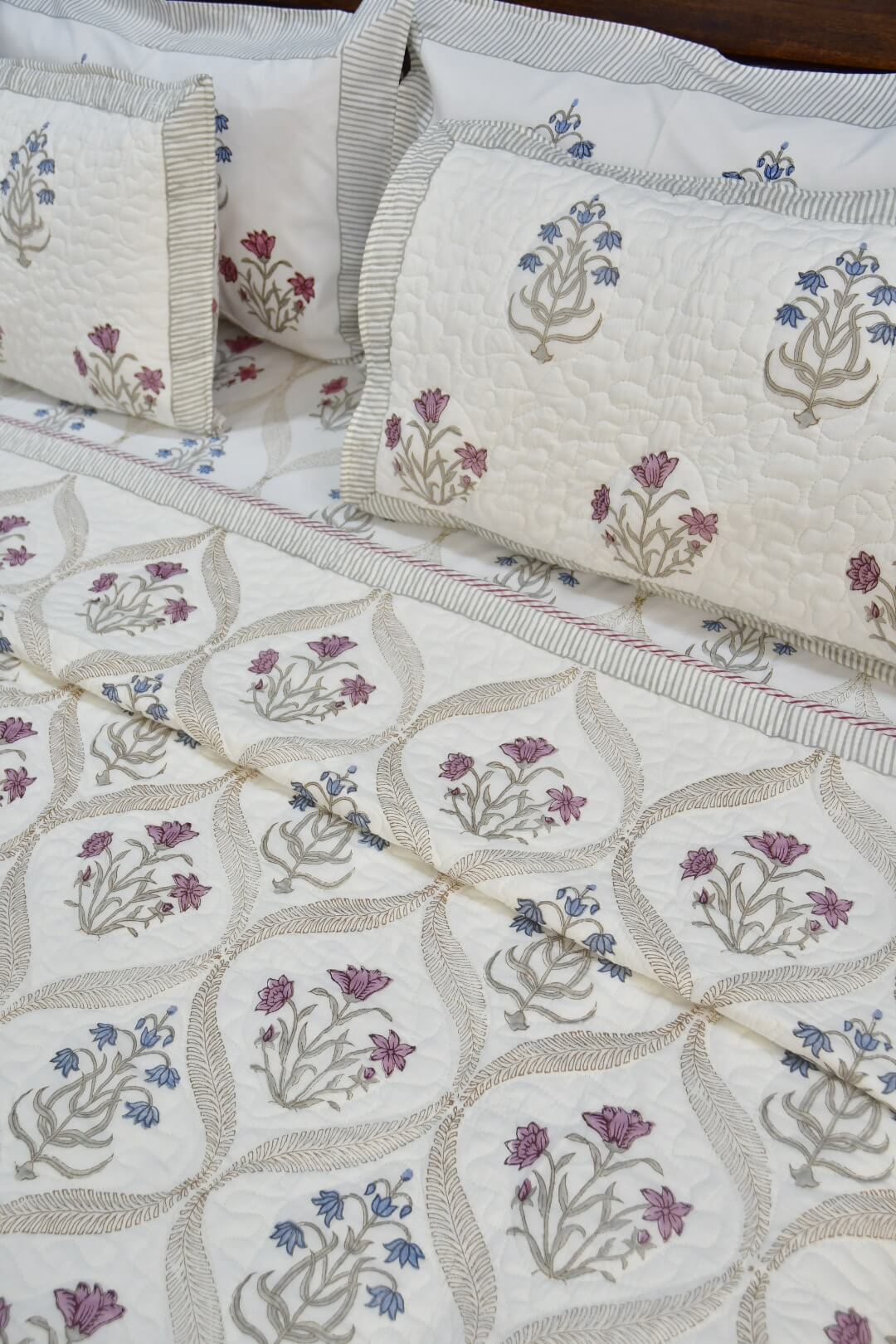 A close-up of a quilted bedding set featuring a cream-colored fabric with a geometric leaf pattern and embroidered floral motifs in purple, blue, and beige, complete with matching pillowcases.