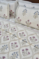 A close-up of a quilted bedding set featuring a cream-colored fabric with a geometric leaf pattern and embroidered floral motifs in purple, blue, and beige, complete with matching pillowcases.