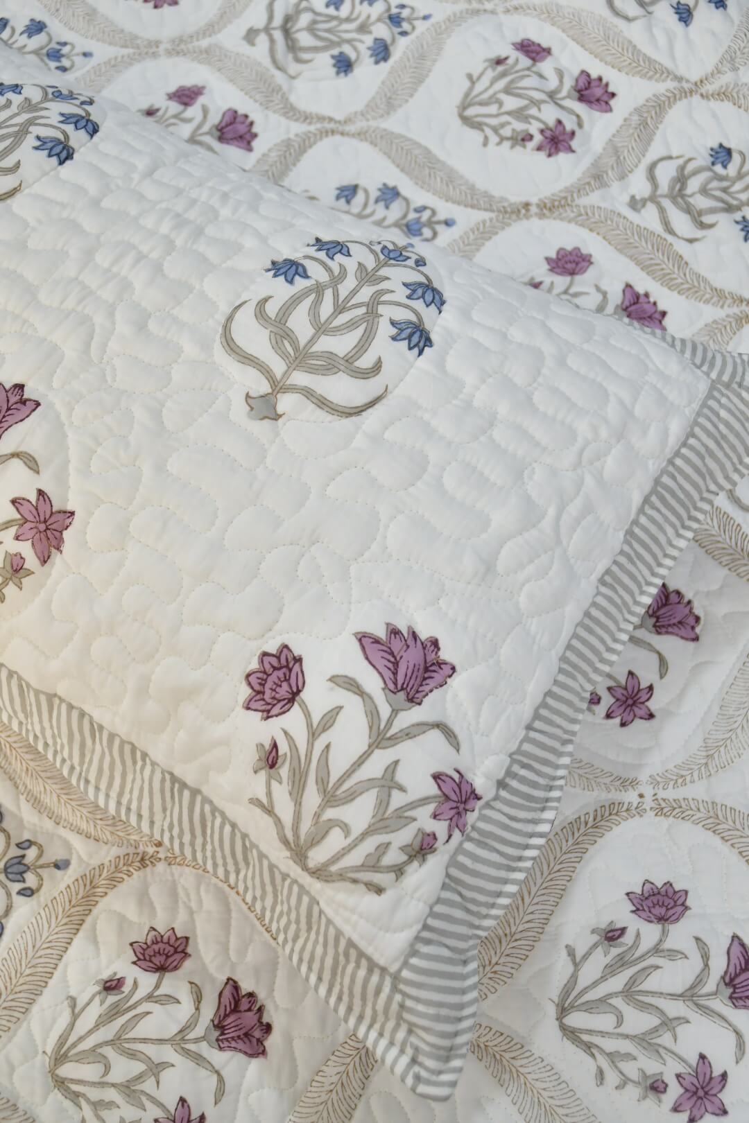 A close-up of a quilted textile product, likely a quilt or comforter, featuring a cream base with embroidered floral motifs in purple, blue, and beige, accented by a striped border.
