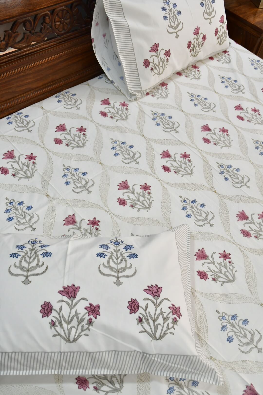 A close-up of a white quilted bedspread with a repeating block-print floral pattern in dusty pink, blue, and gray, styled on a bed with matching pillowcases.