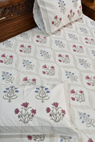 A close-up of a white quilted bedspread with a repeating block-print floral pattern in dusty pink, blue, and gray, styled on a bed with matching pillowcases.