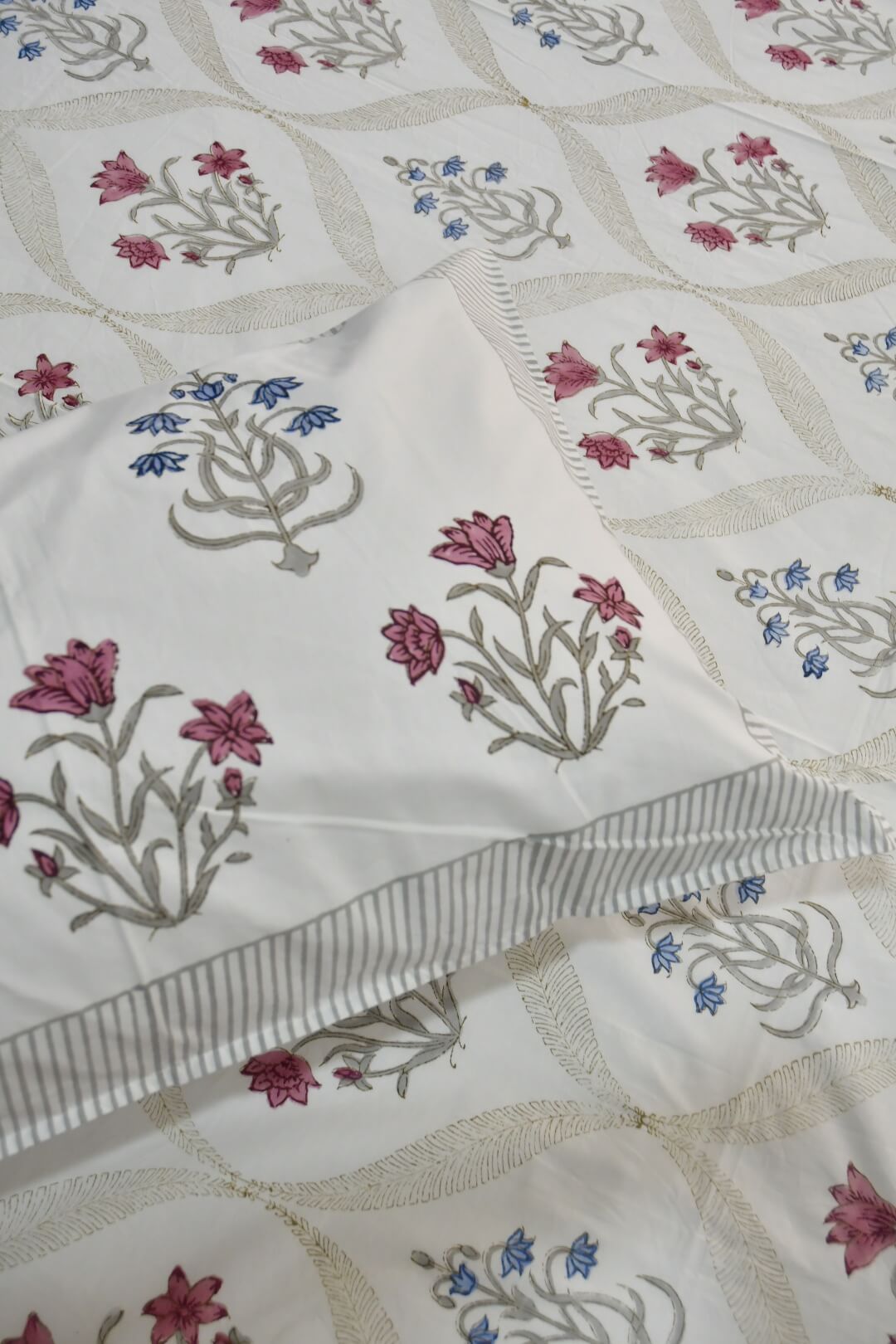 A close-up of a white quilted bedspread or comforter featuring a repeating pattern of embroidered floral motifs in pink, blue, and gray, with a striped border detail.
