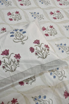 A close-up of a white quilted bedspread or comforter featuring a repeating pattern of embroidered floral motifs in pink, blue, and gray, with a striped border detail.
