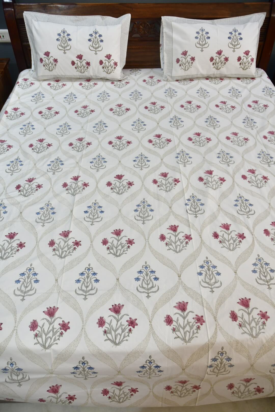 A bedsheet with a cream-colored sheet featuring a repeating floral block print pattern in muted pink, blue, and gray, with two matching pillowcases at the head.