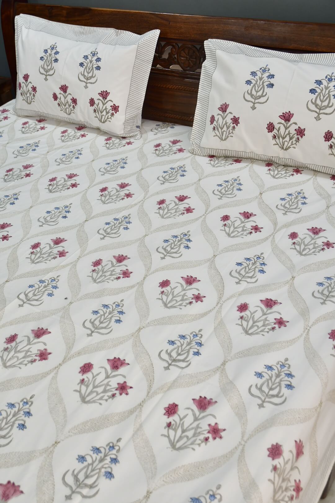 An off-white cotton bedsheet and matching pillowcases featuring a traditional Indian block-print floral pattern in pink, blue, and gray, styled on a bed with a dark wooden headboard.