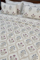 An off-white quilted bedspread with a repeating floral block-print pattern in purple, blue, and beige, styled with matching pillowcases on a bed.