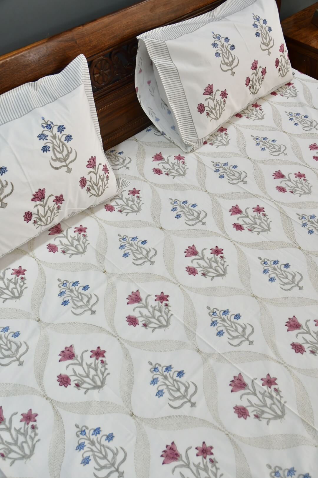 A close-up of a quilted bedspread and matching pillowcases featuring a cream background with a repeating block-print style floral pattern in muted pink, blue, and gray, styled on a bed with a dark wood headboard.