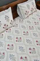 A close-up of a quilted bedspread and matching pillowcases featuring a cream background with a repeating block-print style floral pattern in muted pink, blue, and gray, styled on a bed with a dark wood headboard.