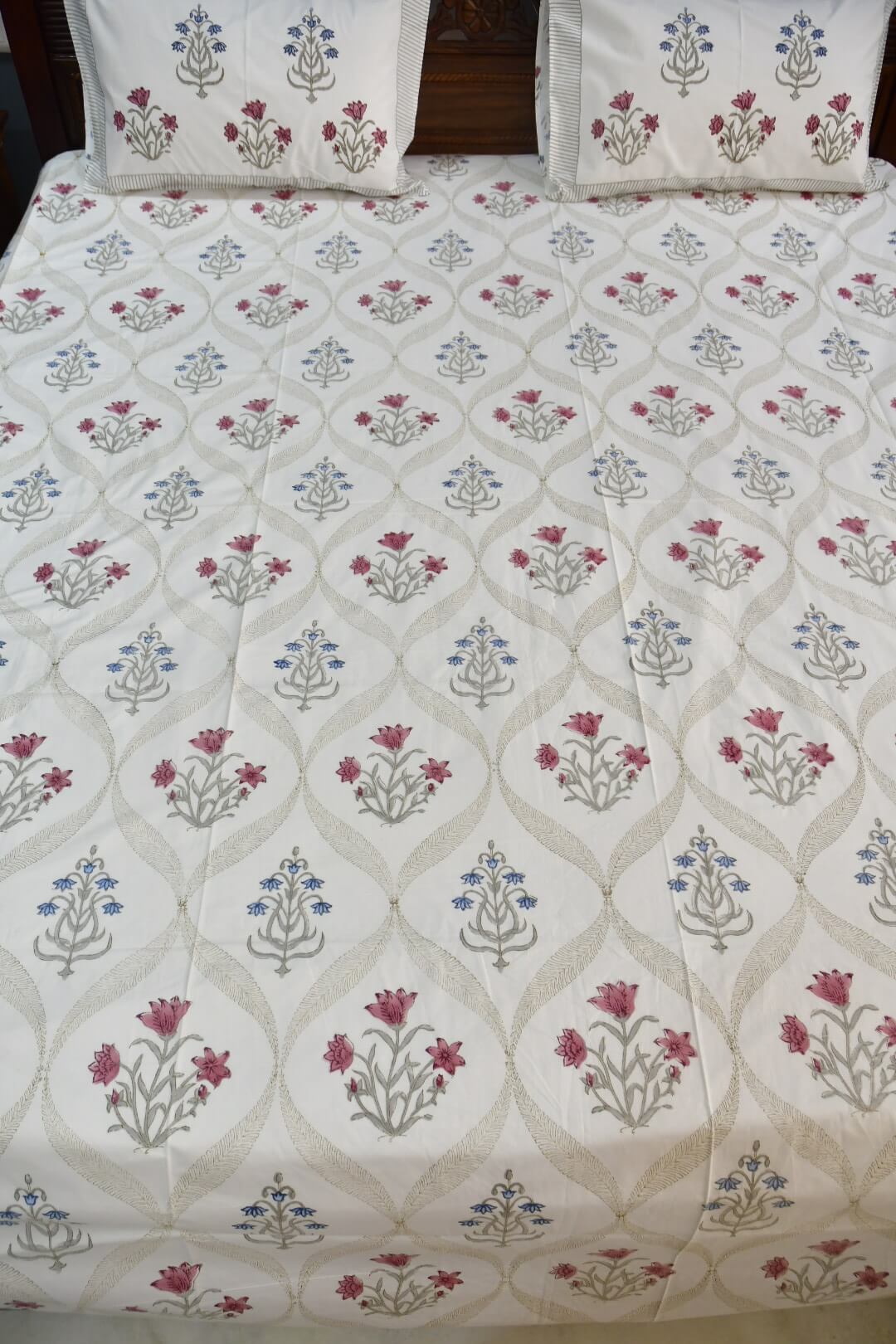 A close-up of a bed dressed with a white quilted comforter or sheet featuring a repeating floral and geometric pattern in muted pink, blue, and gray, with matching pillowcases visible at the top.