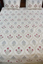 A close-up of a bed dressed with a white quilted comforter or sheet featuring a repeating floral and geometric pattern in muted pink, blue, and gray, with matching pillowcases visible at the top.