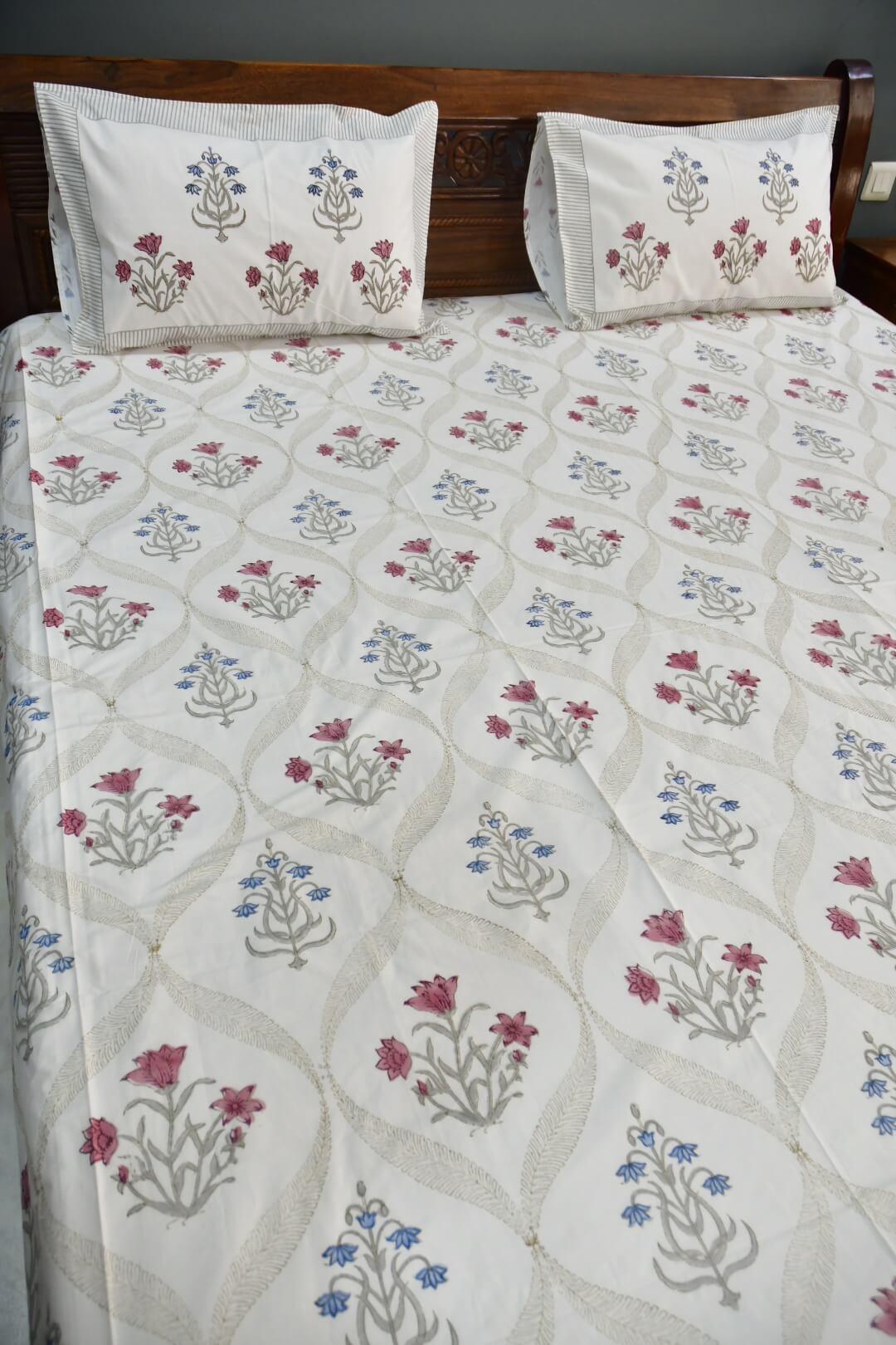 A bed dressed in a white quilted bedspread and matching pillowcases, both decorated with a repeating floral pattern in dusty pink, blue, and gray. The bed is set against a dark brown wooden headboard.
