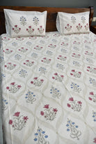 A bed dressed in a white quilted bedspread and matching pillowcases, both decorated with a repeating floral pattern in dusty pink, blue, and gray. The bed is set against a dark brown wooden headboard.