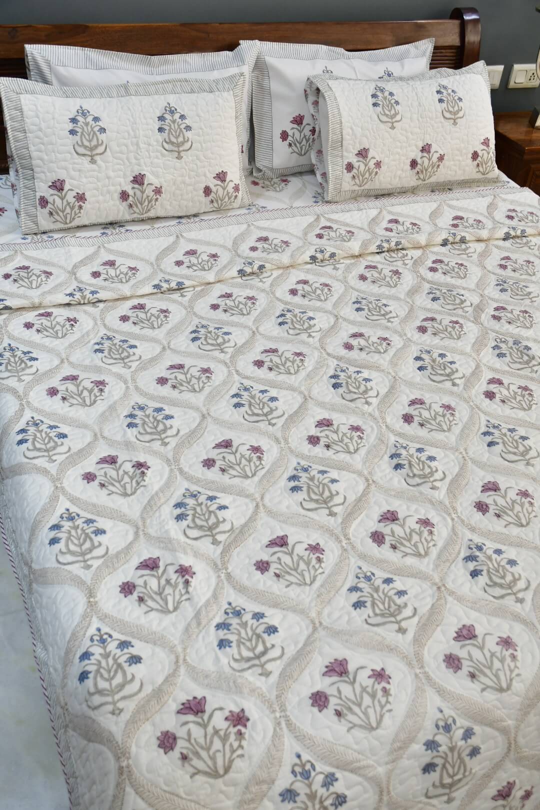 An embroidered quilted bedspread with matching pillowcases, featuring a cream background with beige geometric diamond patterns and blue and purple floral motifs, styled on a bed with a wooden headboard.