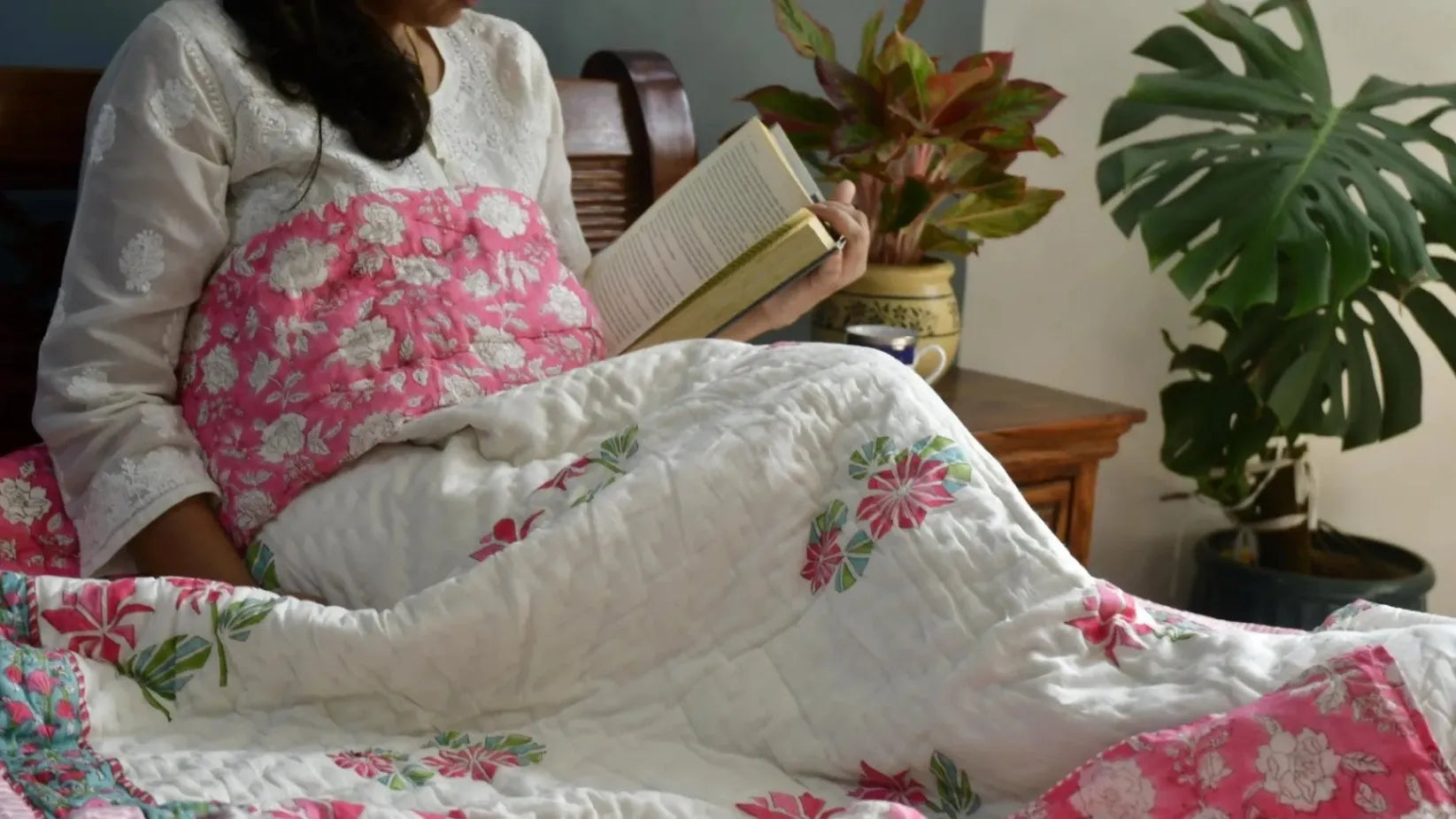 Person reading a book on a floral quilt with plants in the background