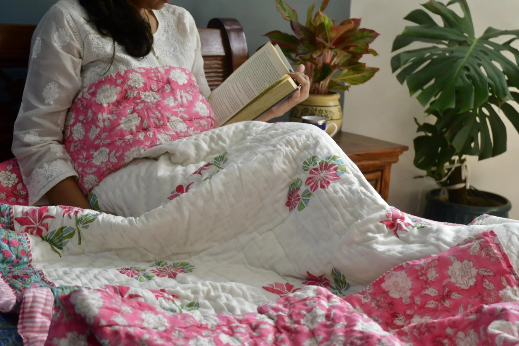 Person sitting on a couch under a floral quilt, reading a book in a cozy room with plants.