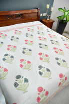 A white dohar / AC comforter with a repeating floral block-print pattern in pink, gray, and green, styled on a bed with a dark wood headboard in a bedroom setting.