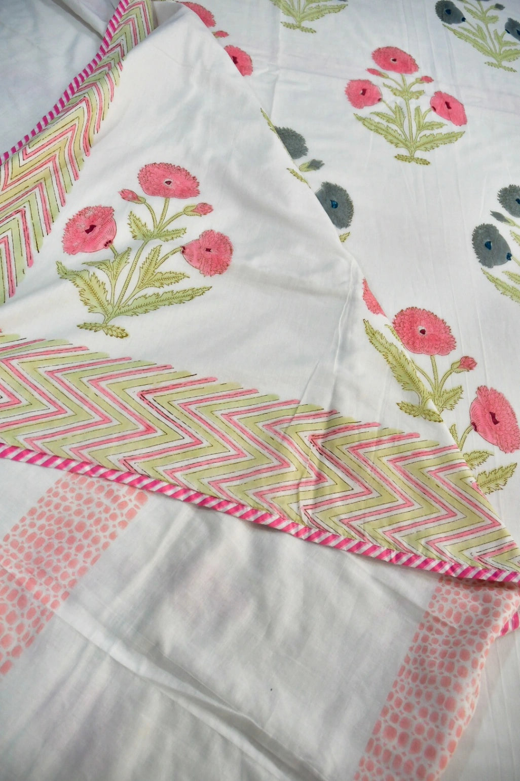 White fabric textile with pink floral prints, green stems, and pink-and-green chevron border details, laid out in a close-up view.
