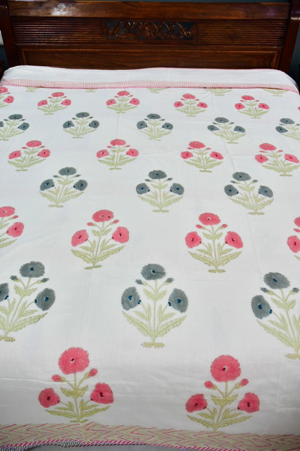 A white cotton bedspread or dohar featuring a repeating block-print style floral pattern in pink, gray, and green, with a pink and white striped border.