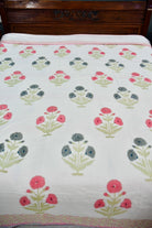 A white cotton bedspread or dohar featuring a repeating block-print style floral pattern in pink, gray, and green, with a pink and white striped border.