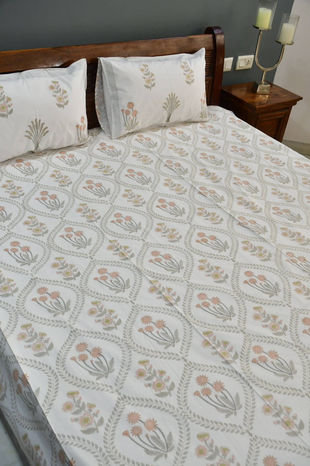 A bed dressed in a white printed sheet and matching pillowcases featuring a repeating floral and geometric pattern in soft beige, gray, and muted orange tones, shown in a bedroom setting with a wooden headboard and bedside table.