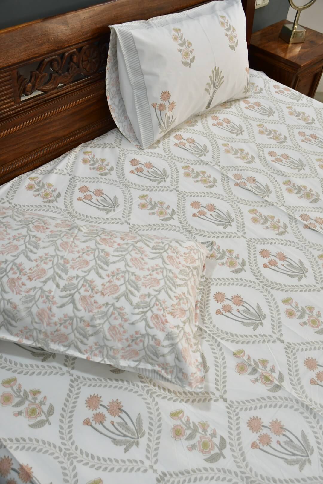 A close-up of a bed dressed in a white printed sheet and pillowcases featuring a repeating floral and geometric motif in soft sage green, blush pink, and muted yellow, with a dark wood headboard and bedside table visible.