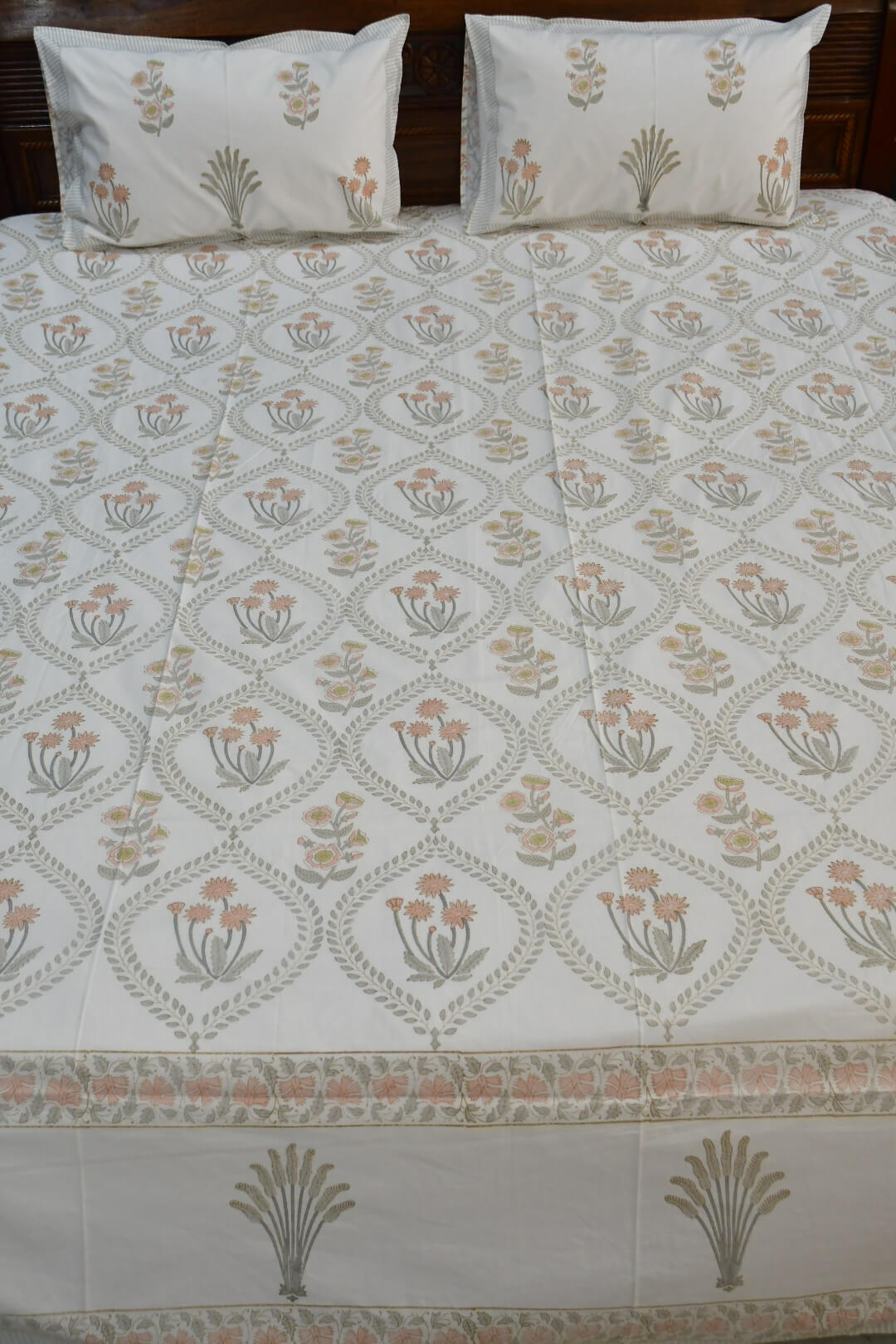 A bed dressed with a white cotton bedsheet featuring a repeating peach and gray floral block print, with two matching pillowcases resting against a dark wooden headboard.