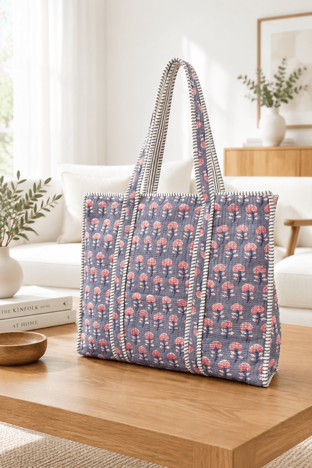A large woven tote bag with a blue-gray base and pink floral pattern, featuring striped handles, displayed on a wooden table in a bright, modern living room.