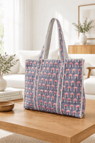 A large woven tote bag with a blue-gray base and pink floral pattern, featuring striped handles, displayed on a wooden table in a bright, modern living room.