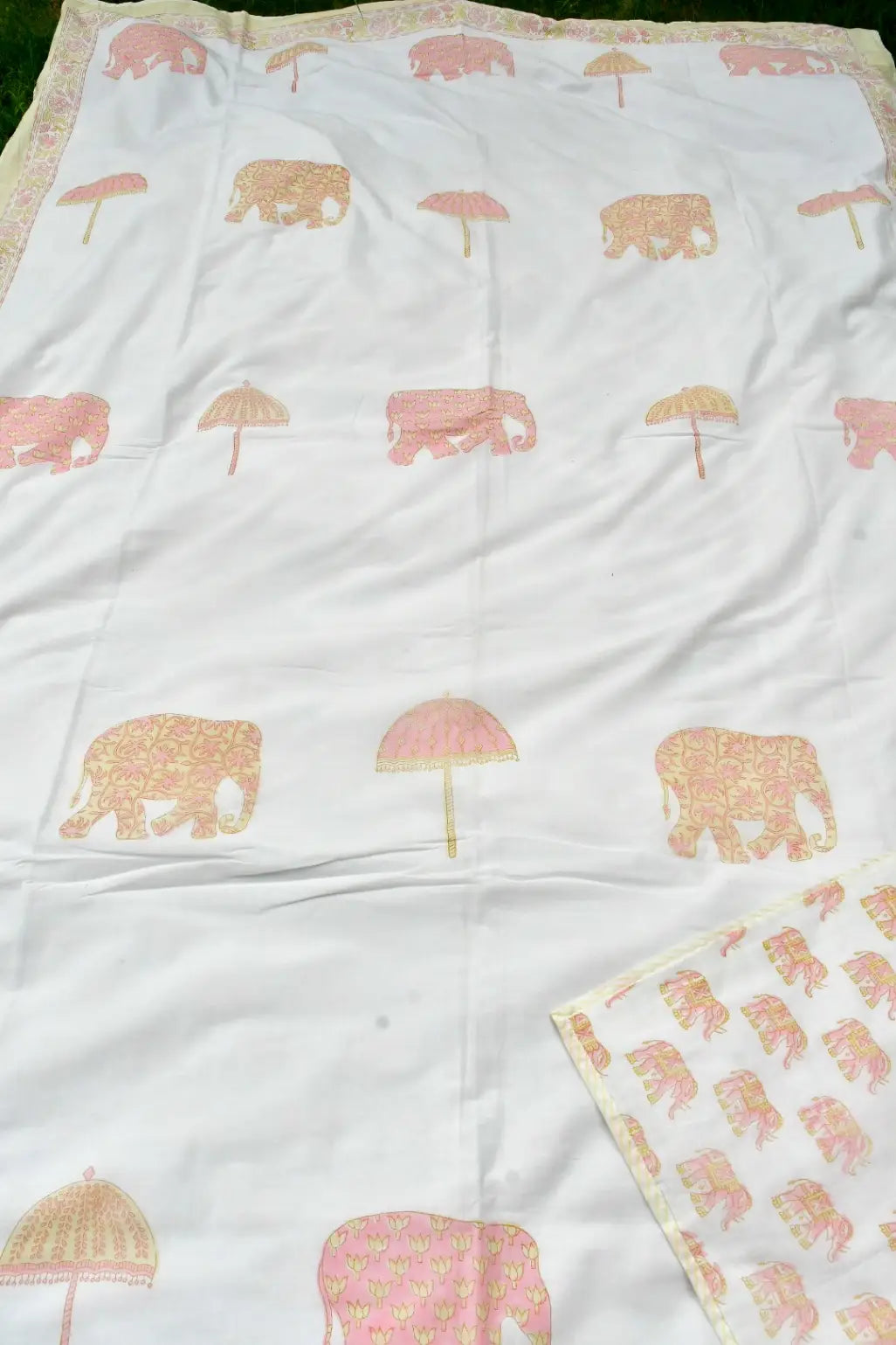 A white dohar  featuring a repeating pattern of pink elephants and pink umbrellas with gold floral detailing, laid flat with a matching patterned fabric piece folded in the bottom right corner.