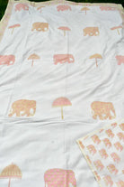 A white dohar  featuring a repeating pattern of pink elephants and pink umbrellas with gold floral detailing, laid flat with a matching patterned fabric piece folded in the bottom right corner.