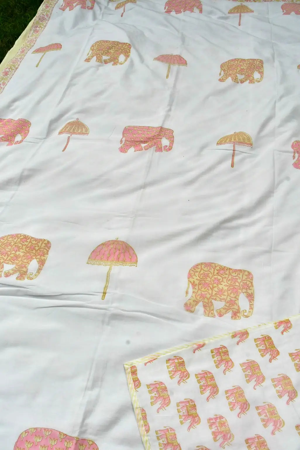 A white dohar featuring a repeating block-print pattern of pink elephants and pink umbrellas with gold detailing, laid out on a green surface.