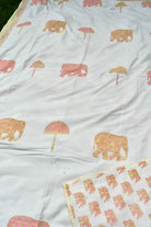A white dohar featuring a repeating block-print pattern of pink elephants and pink umbrellas with gold detailing, laid out on a green surface.