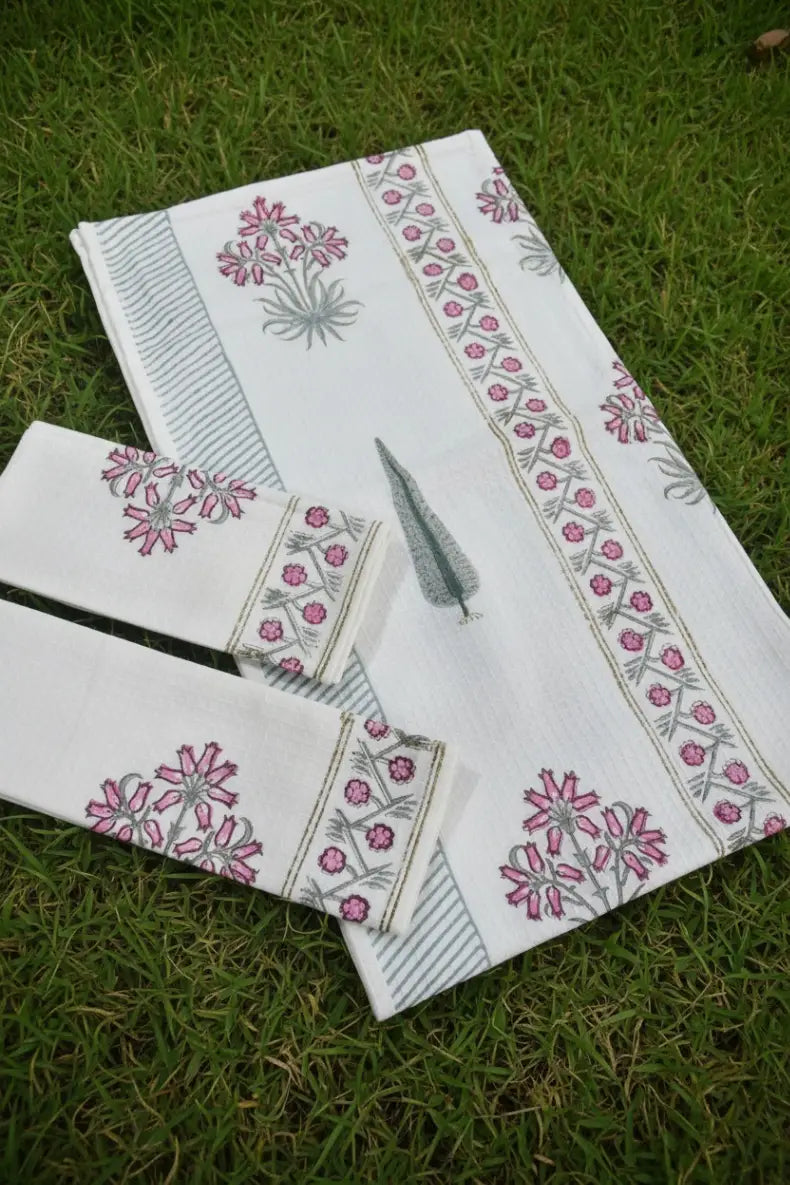 A set of white cotton towel sets, featuring pink and gray floral block prints and borders, displayed on a green grass background.