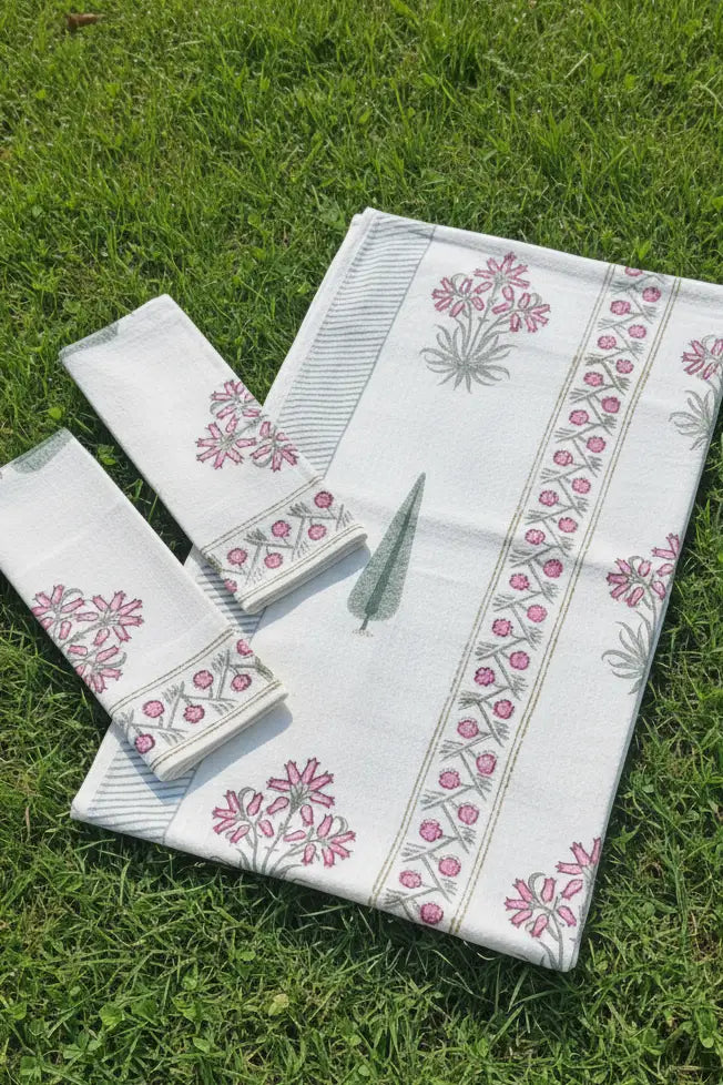 A white cotton towel set with pink and green floral block prints, laid out on a green grass lawn. The set includes a rectangular cloth, a matching square cloth, and a smaller rectangular cloth, all featuring striped borders.