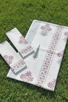 A white cotton towel set with pink and green floral block prints, laid out on a green grass lawn. The set includes a rectangular cloth, a matching square cloth, and a smaller rectangular cloth, all featuring striped borders.