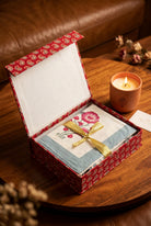 A folded embroidered cloth set inside an open patterned gift box, tied with a gold ribbon. The cloth has a light blue border, a white center with pink floral embroidery, and red heart accents.