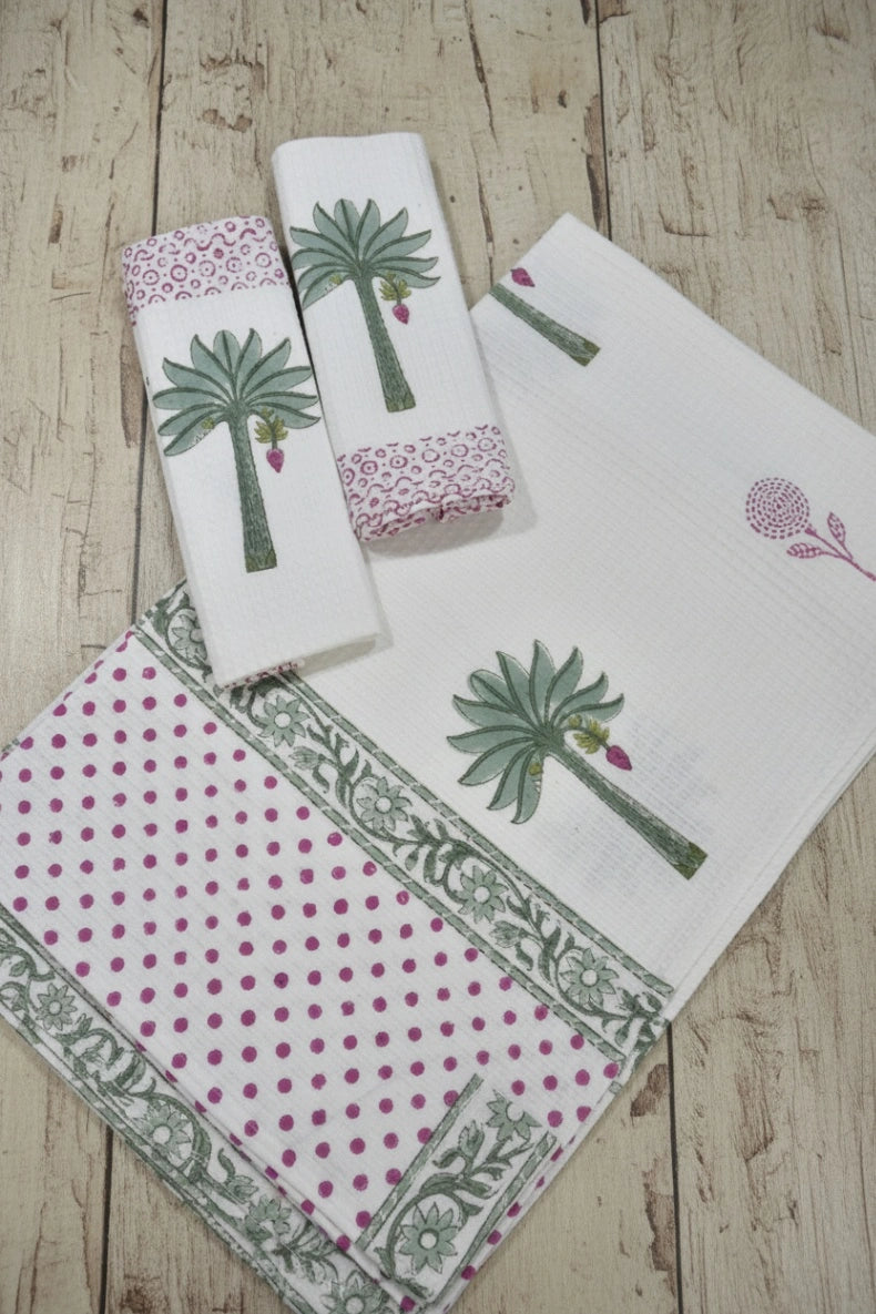 A set of white cotton kitchen towels and a matching napkin featuring green palm tree prints, pink and green polka dots, and ornate borders, arranged on a light wood background.